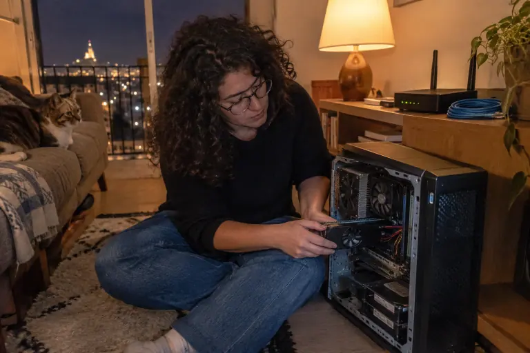 Une femme installe une petite carte graphique demi-hauteur à un ventilateur dans un serveur domestique ouvert, le soir, à côté d’un routeur