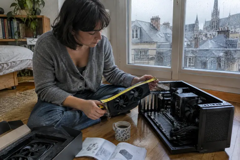 À Rouen, une femme assise sur le parquet mesure au mètre ruban une carte graphique à deux ventilateurs à côté d’un petit boîtier ouvert