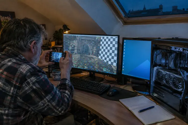 Un homme barbu photographie avec son téléphone un écran de jeu figé couvert de points colorés et d’un motif en damier, dans un bureau mansardé