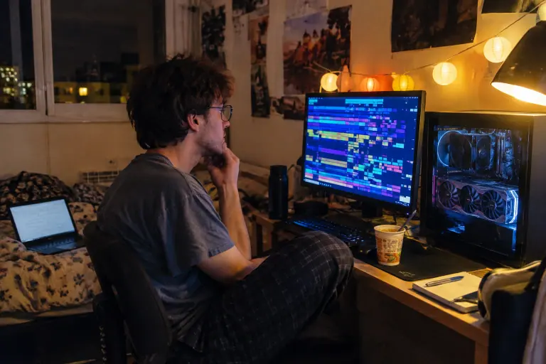 Un étudiant observe tard le soir l’écran de son PC à grosse carte graphique dans sa chambre de résidence à Toulouse, ses notes de cours ouvertes sur le lit