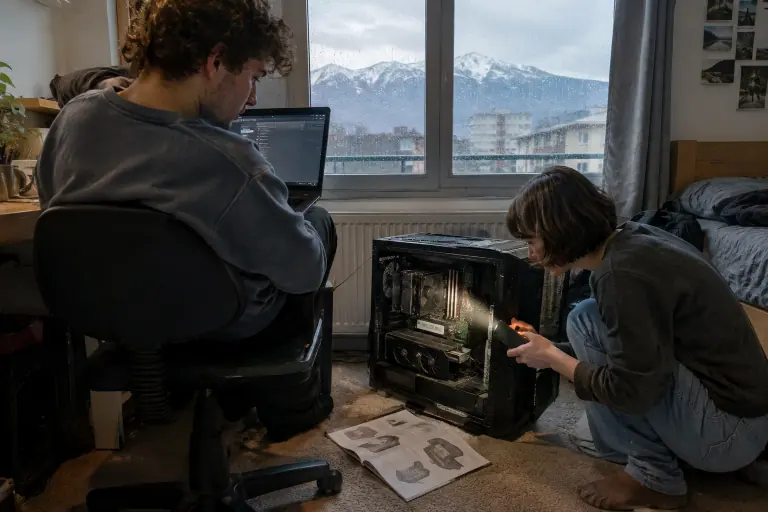 Dans une chambre d’étudiant, une jeune femme éclaire la carte graphique d’un PC ouvert pendant que son colocataire consulte un portable