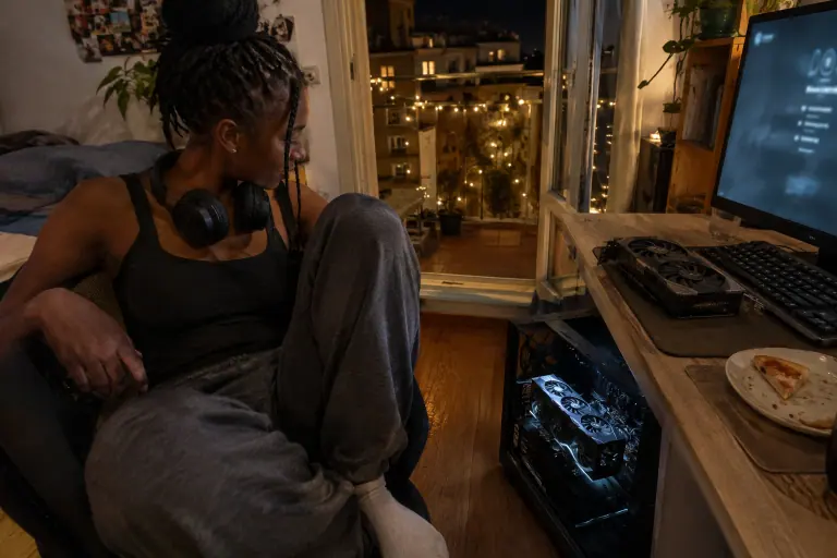 Tard le soir, une jeune femme au casque autour du cou regarde la grande carte graphique de son PC posé au sol, une ancienne carte sur le bureau