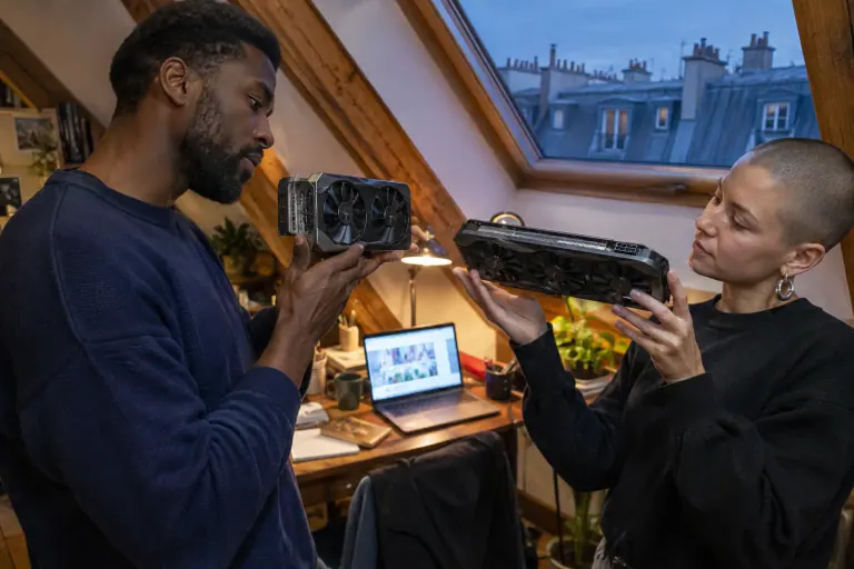 Sous les toits à Paris, deux amis comparent côte à côte l’épaisseur d’une carte graphique à deux ventilateurs et d’une autre à trois ventilateurs