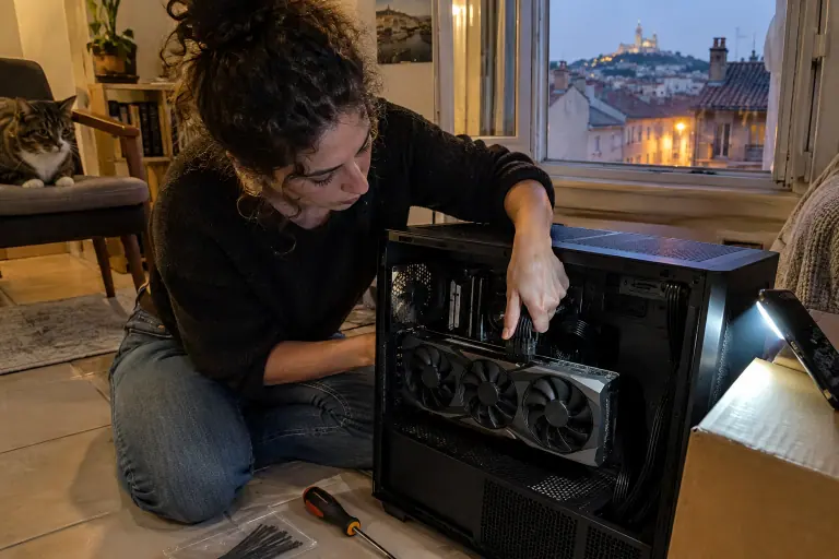 Dans un appartement à Marseille, une femme enfonce le câble 16 broches dans une grande carte graphique à trois ventilateurs, éclairée par son téléphone