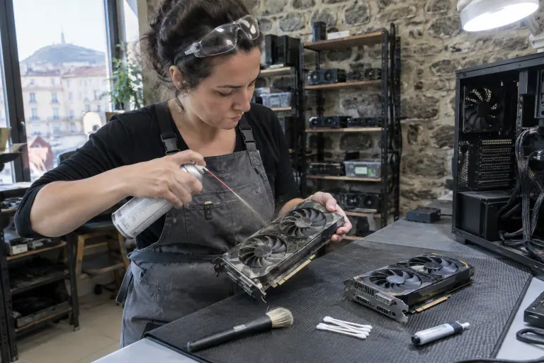 Dans un atelier de réparation à Clermont-Ferrand, une technicienne dépoussière à la bombe d’air une carte graphique d’occasion à deux ventilateurs