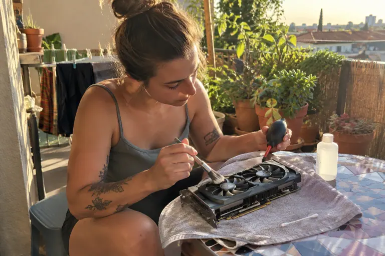 Sur un balcon ensoleillé, une jeune femme dépoussière au pinceau une carte graphique d’occasion à deux ventilateurs posée sur une serviette