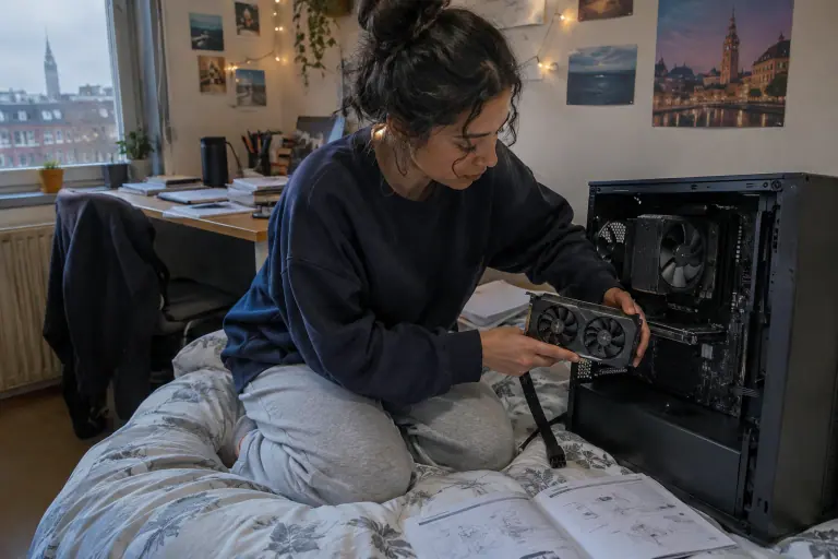 Une étudiante installe une carte graphique compacte à deux ventilateurs dans un petit boîtier ouvert posé sur son lit, notice de carte mère à côté