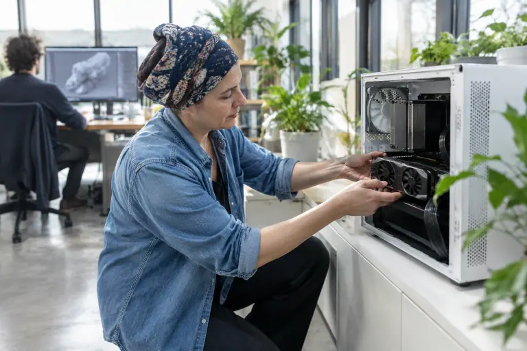Une graphiste 3D insère une fine carte graphique à deux ventilateurs dans un petit PC blanc, dans un studio de création lumineux à Nantes