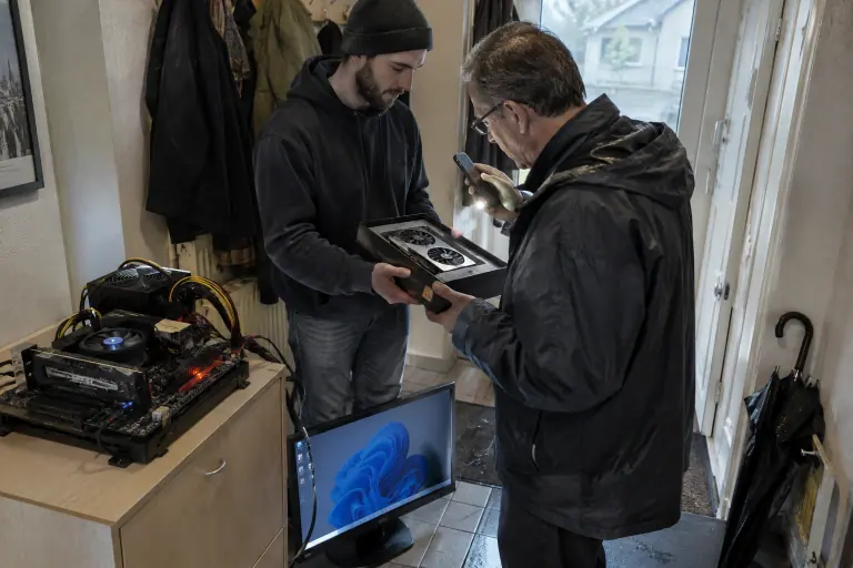 Dans l’entrée d’un appartement, un jeune homme tend sa carte graphique dans sa boîte à un acheteur qui l’inspecte, un banc de test installé à côté