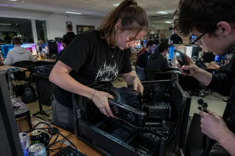 Lors d’une LAN party dans une salle des fêtes, une adolescente installe une carte graphique à deux ventilateurs pendant qu’un ami l’éclaire