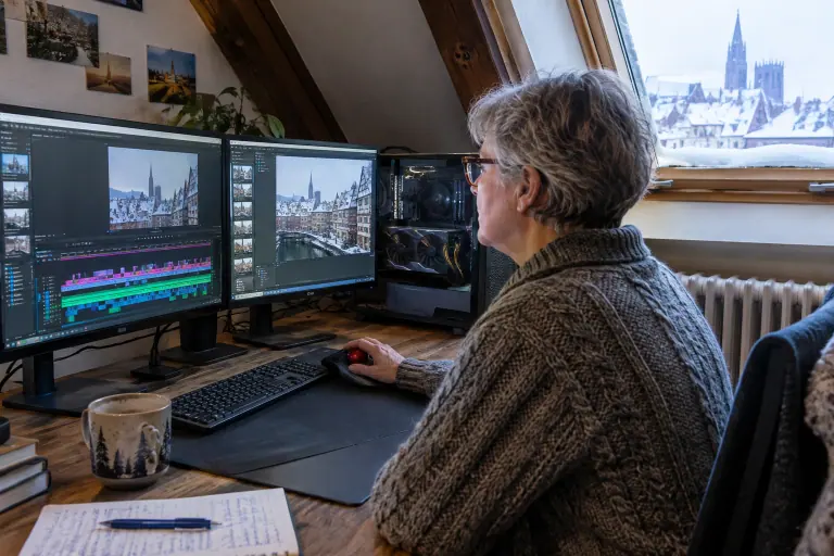 Une femme monte une vidéo familiale sur deux écrans dans un bureau mansardé, à côté d’une tour vitrée équipée d’une longue carte graphique