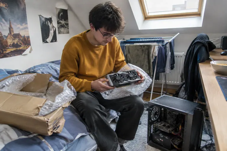 Un étudiant assis sur son lit déballe du papier bulle une carte graphique d’occasion à deux ventilateurs, son PC ouvert posé au sol à ses pieds