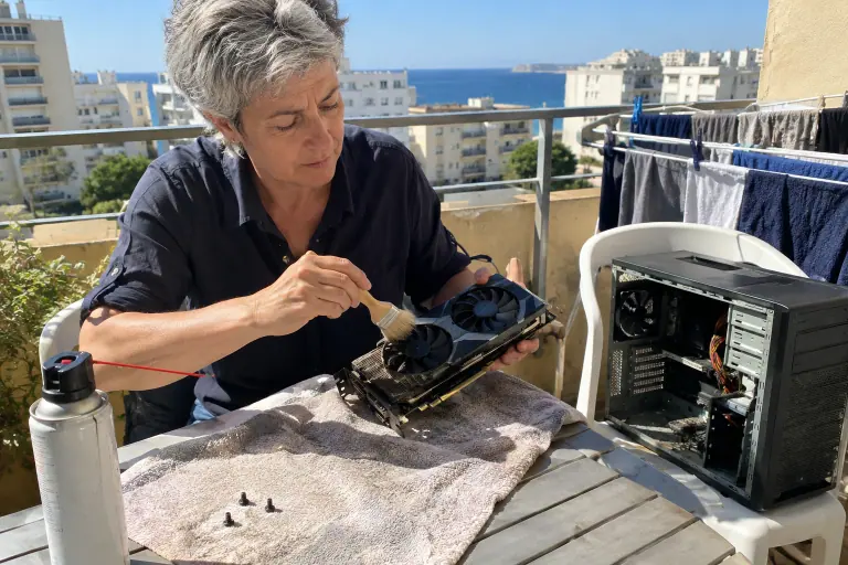 Sur un balcon à Marseille, une femme dépoussière au pinceau les ailettes d’une carte graphique d’occasion à deux ventilateurs