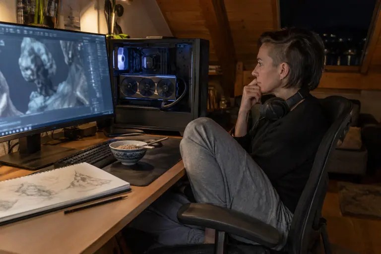 Tard le soir à Annecy, une graphiste 3D suit un rendu sur son grand écran, à côté de son PC ouvert équipé d’une longue carte graphique à trois ventilateurs