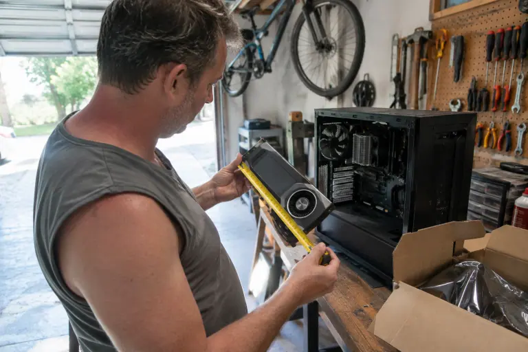 Dans un garage, un homme mesure au mètre ruban la longueur d’une carte graphique à turbine devant une tour PC ouverte posée sur l’établi