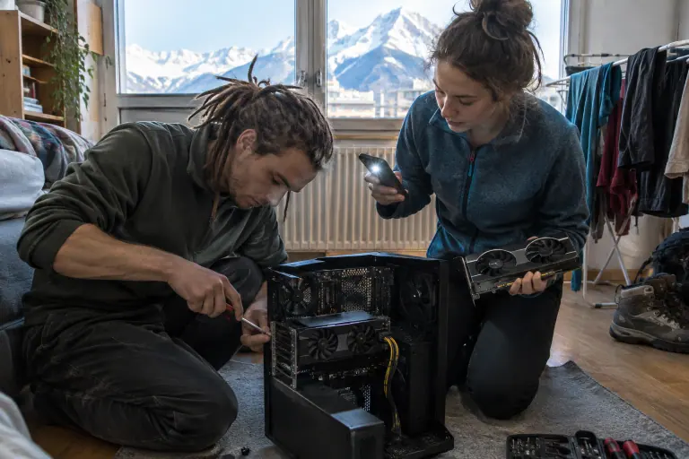 Deux colocataires installent une carte graphique à deux ventilateurs dans une tour posée au sol, devant une fenêtre donnant sur les montagnes