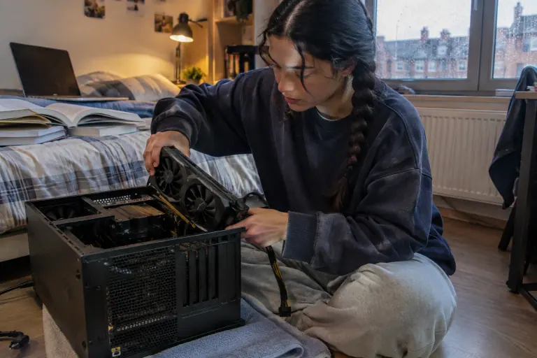 Une étudiante installe une carte graphique compacte à deux ventilateurs dans un petit boîtier micro-ATX posé sur une serviette dans sa chambre