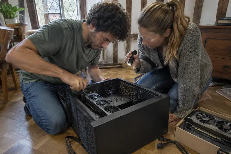 Un homme visse l’équerre d’une longue carte graphique à trois ventilateurs dans une tour couchée, sa compagne éclairant l’intérieur avec une lampe