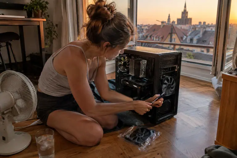 Un soir d’été, une jeune femme visse un ventilateur de 140 mm en façade de son boîtier PC ouvert, posé au sol près d’une fenêtre ouverte